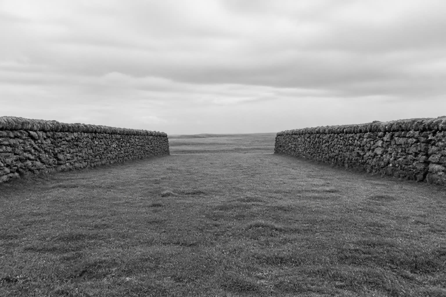 Black-and-white two parallel stone walls that stop short of meeting in an open field